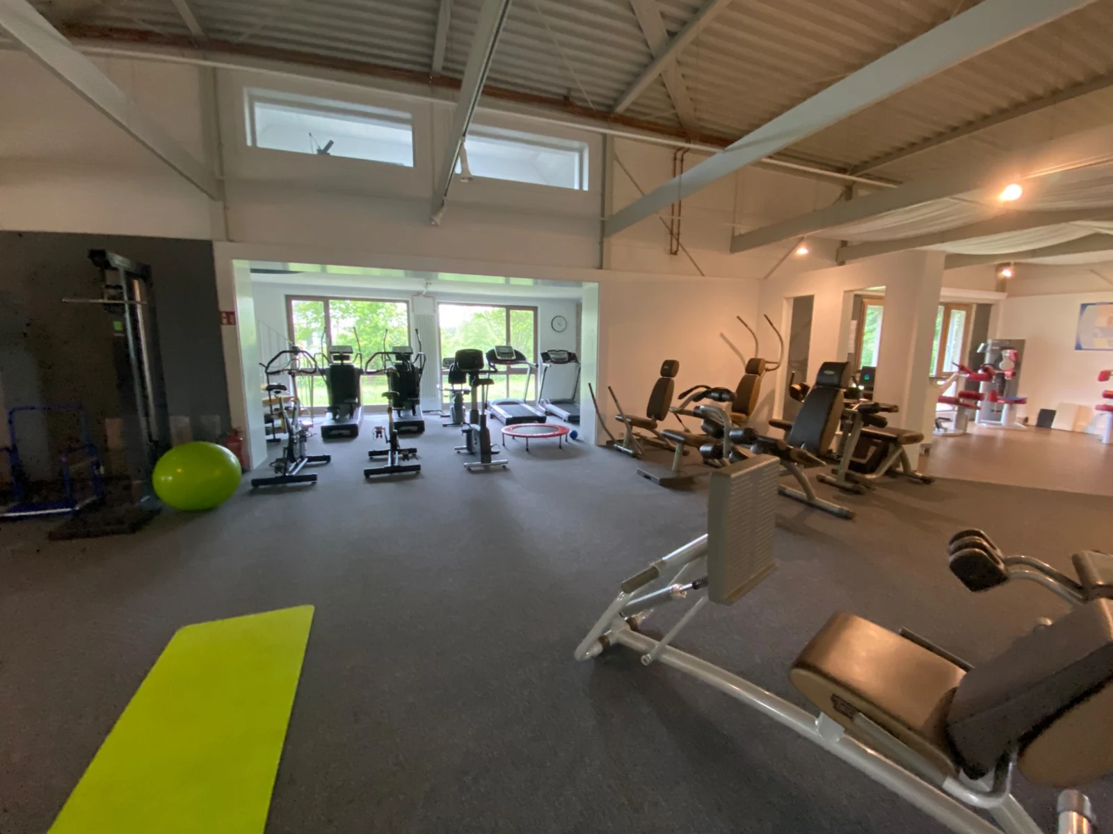 Ausdauergeräte vor der Fensterfront sowie Trainingsgeräte und freie Fläche im Fitnessstudio