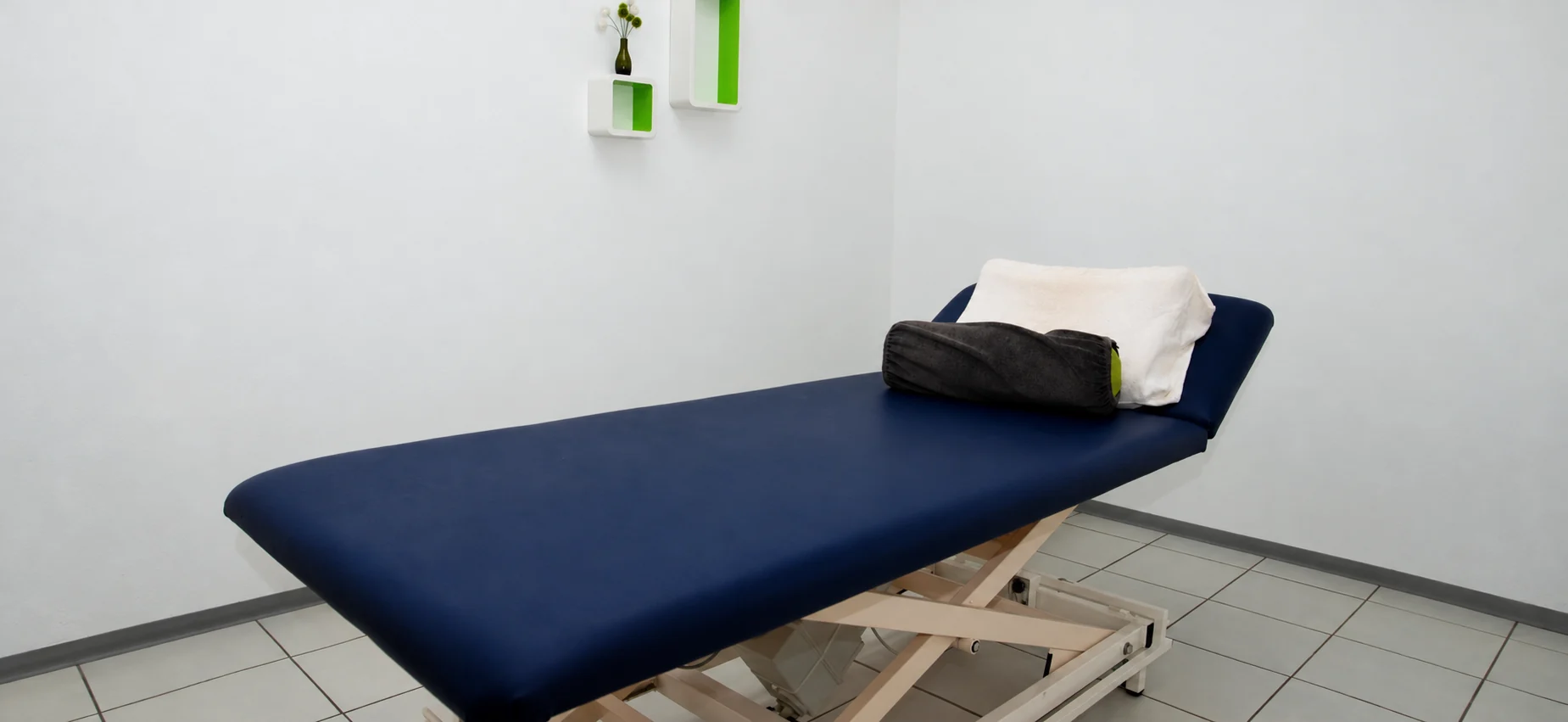 Helle Behandlungskabine mit blauer Therapieliege, weißem Kissen, dunkler Rolle und grünen Wandnischen