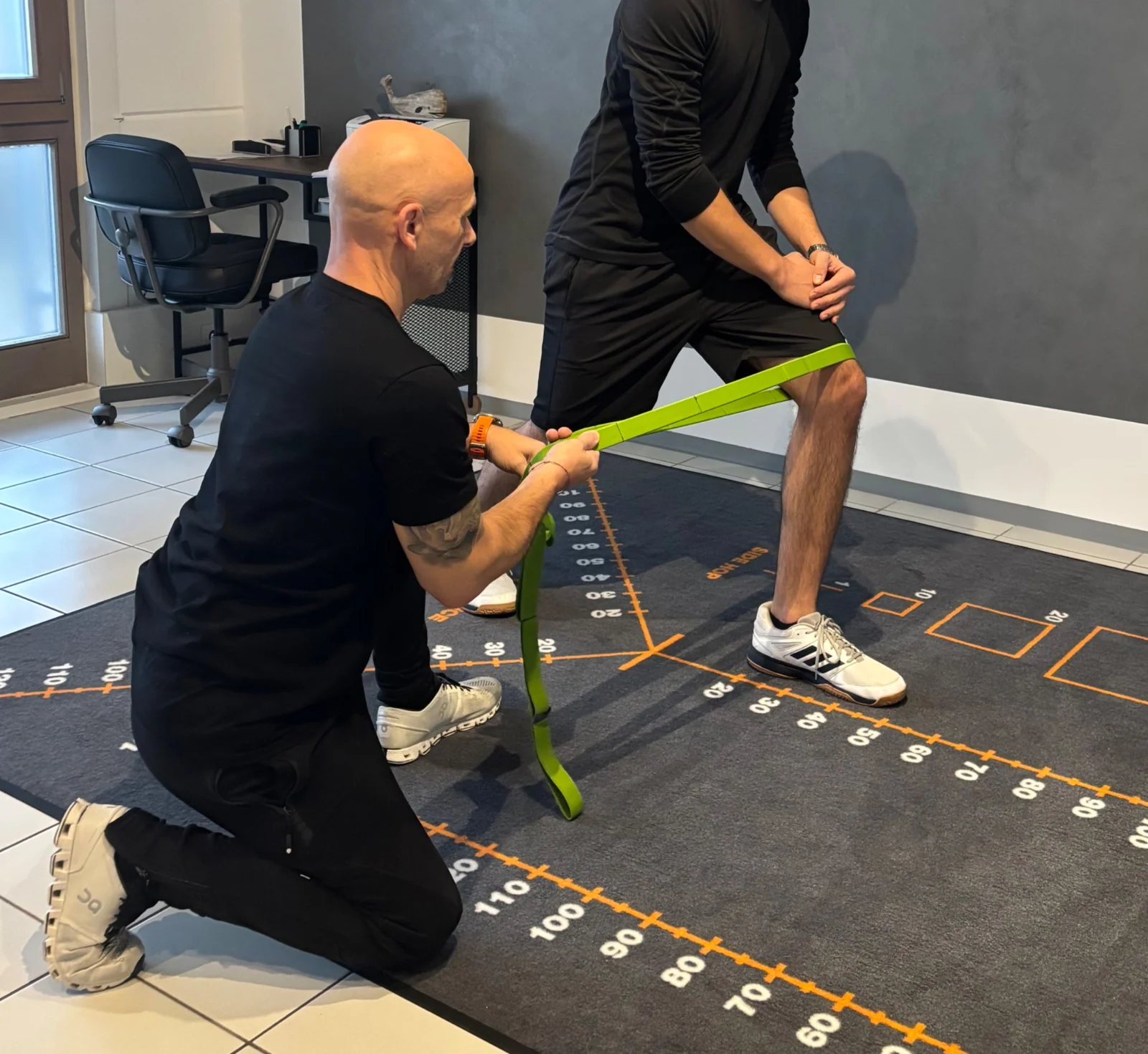 Sportphysiotherapeut begleitet eine Beinübung mit einem grünen Widerstandsband auf einer markierten Trainingsfläche